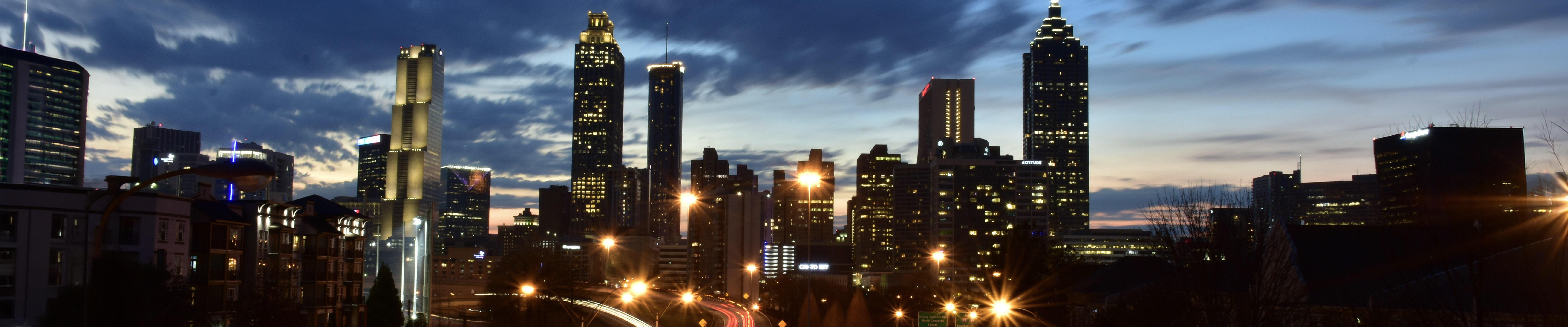 A dusk cityscape shows a modern skyline with tall, illuminated buildings under streaky blue clouds. In the foreground, a multi-lane highway curves into the city, captured with long exposure so headlights form white streams and taillights form red ribbons. Streetlights glow amber along the road, and the horizon still holds a faint band of sunset light. The scene feels dynamic and energetic as traffic flows toward the downtown core.
