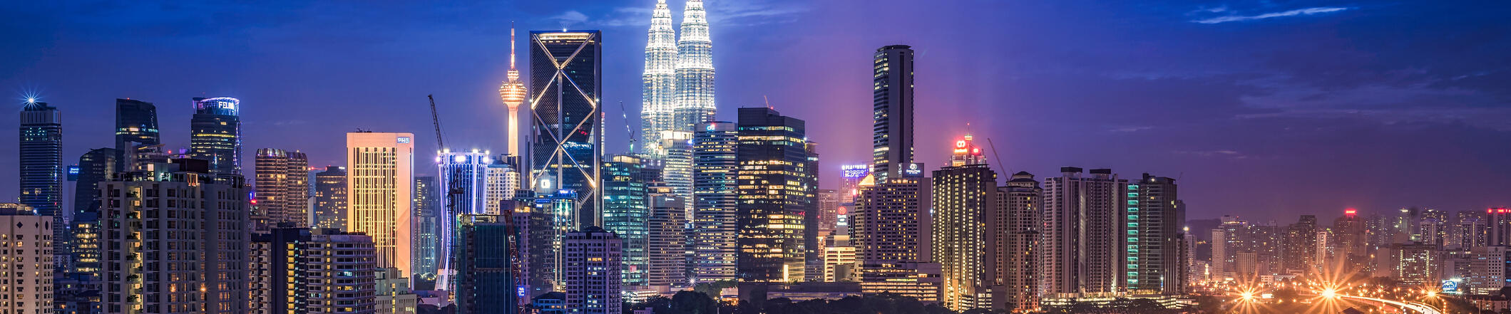 A vibrant city skyline glows at dusk, with numerous high-rise buildings illuminated against a deep blue sky.