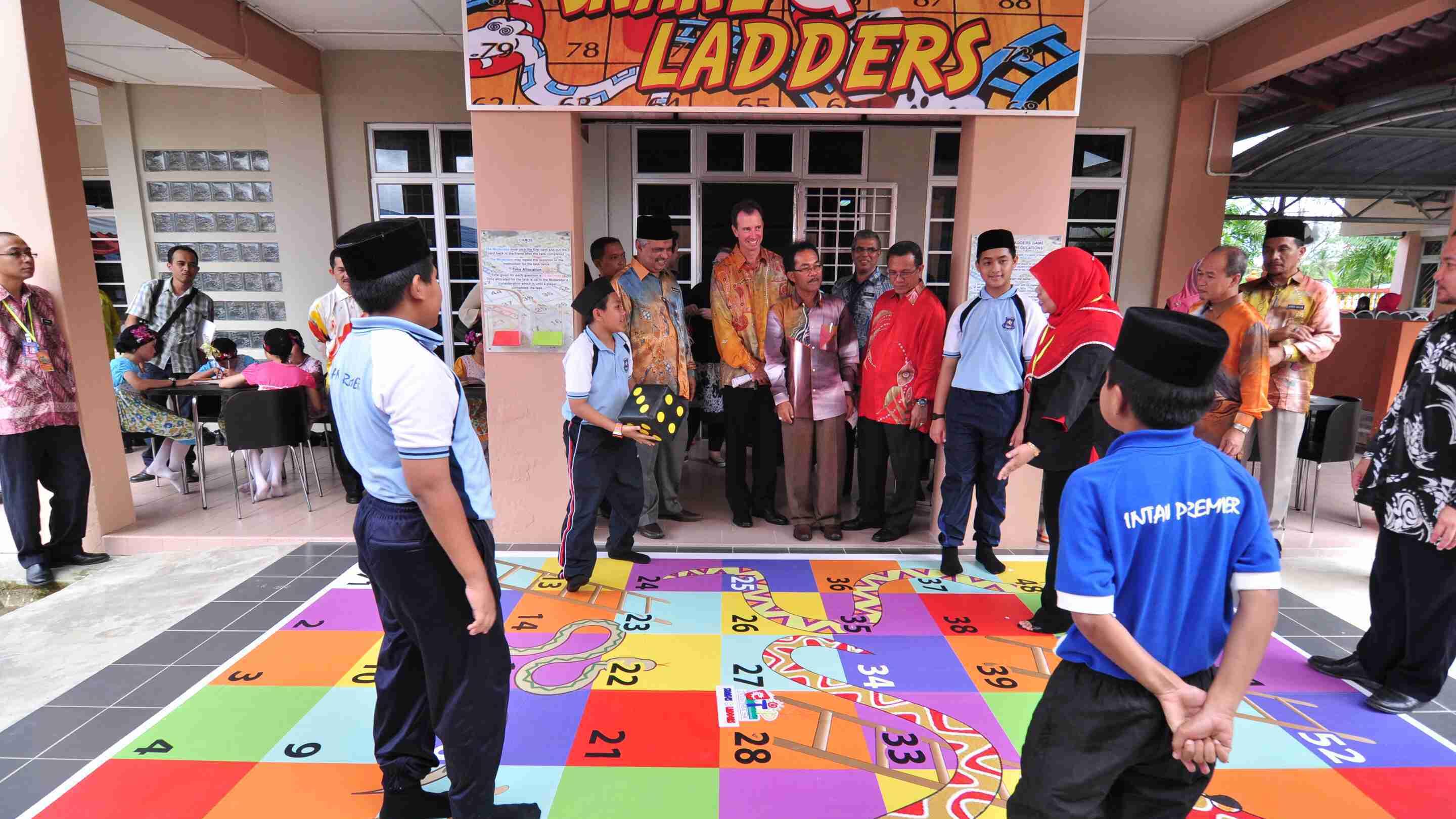 A group of students and adults are gathered around a large floor version of the Snakes and Ladders board game.