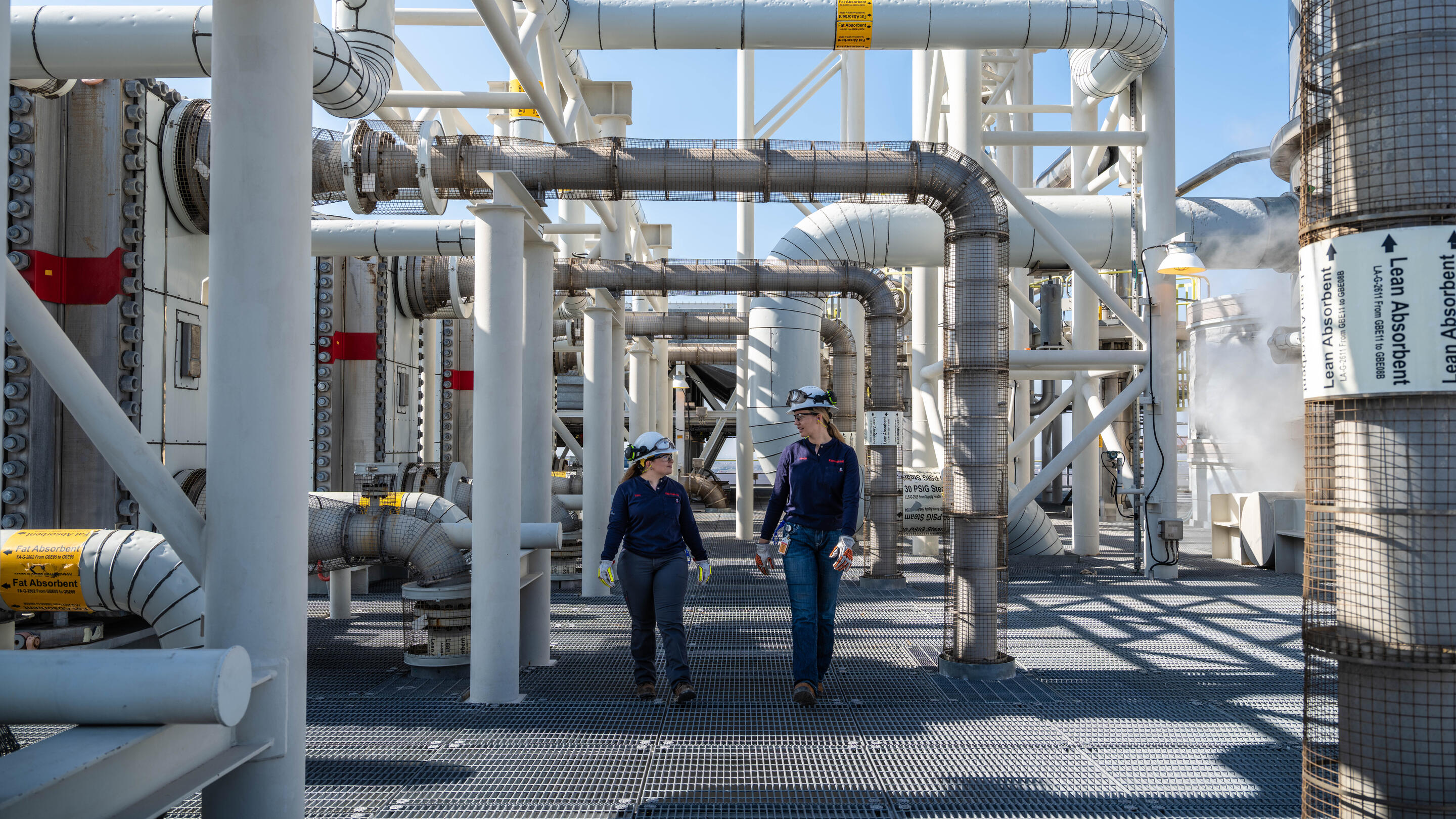 The image shows an industrial facility filled with large white pipes, ducts, and support structures under a clear blue sky. Two workers wearing hard hats, safety glasses, gloves, and blue uniforms walk along a grated metal walkway between the equipment.