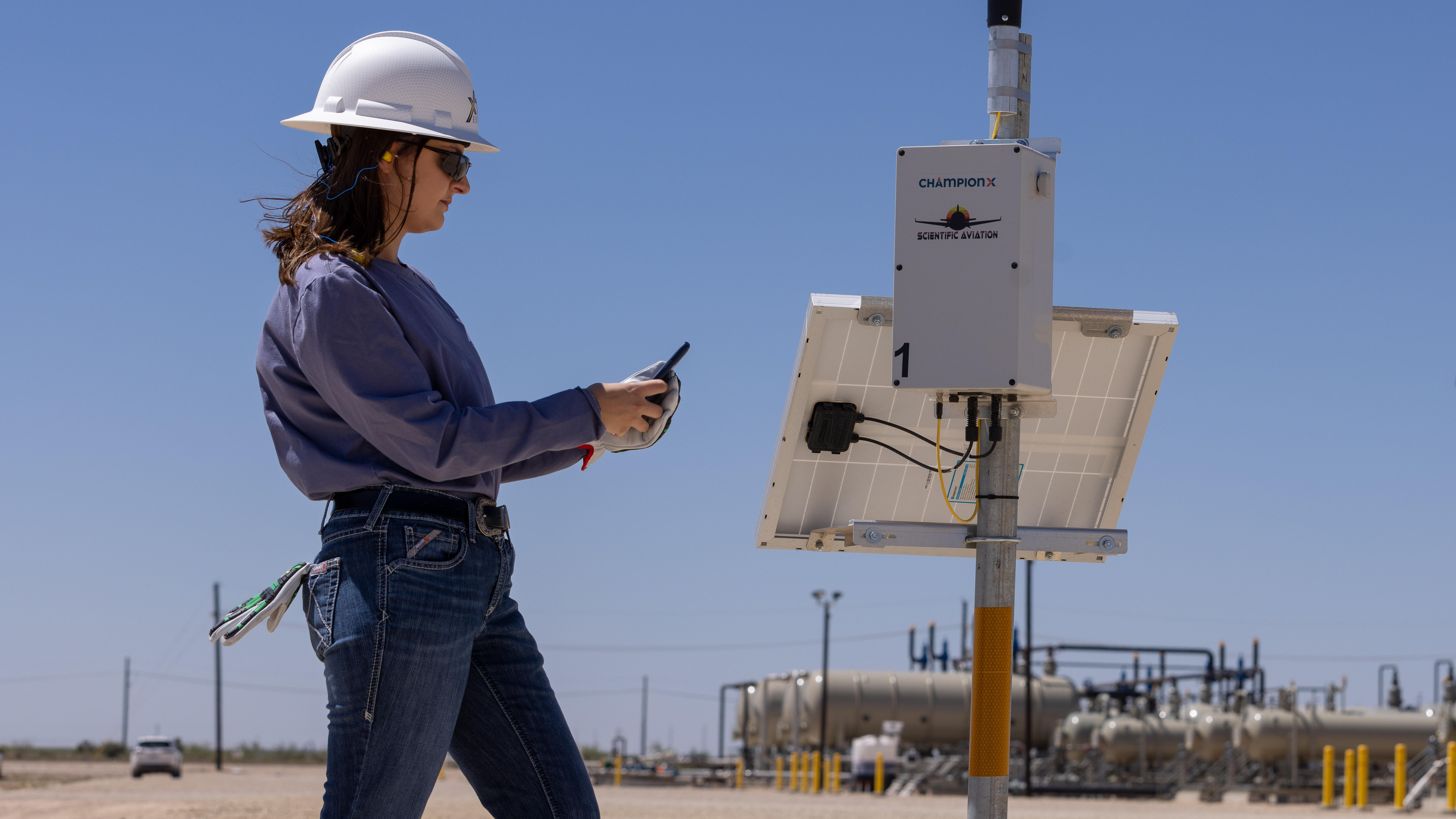 A person wearing a white hard hat, blue long-sleeve shirt, and jeans is standing outdoors at XTO operations in the Permian Basin.